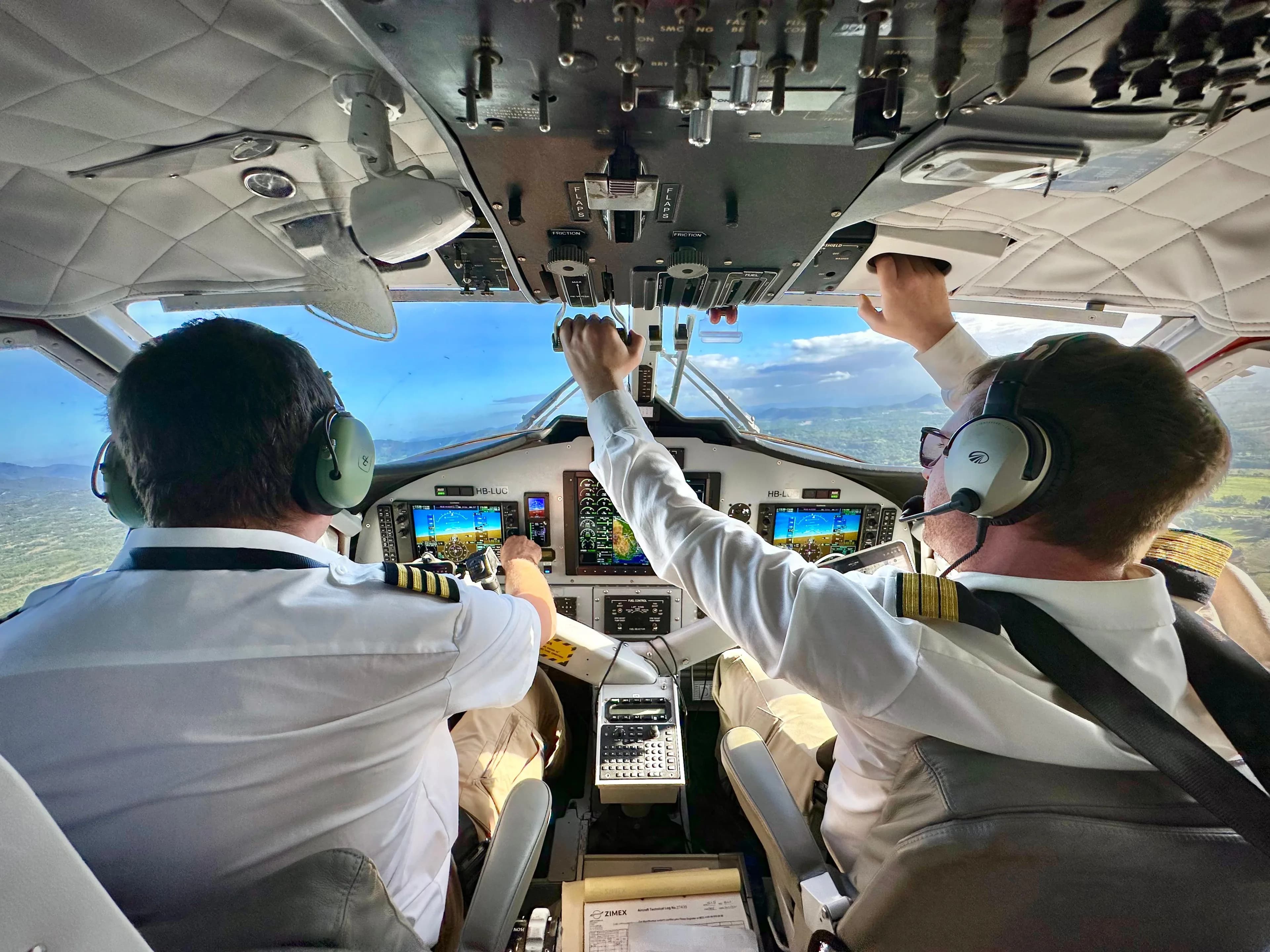 Two Zimex pilots seen from behind on the flight deck of a Twin Otter, one reaching up to the overhead panel.
