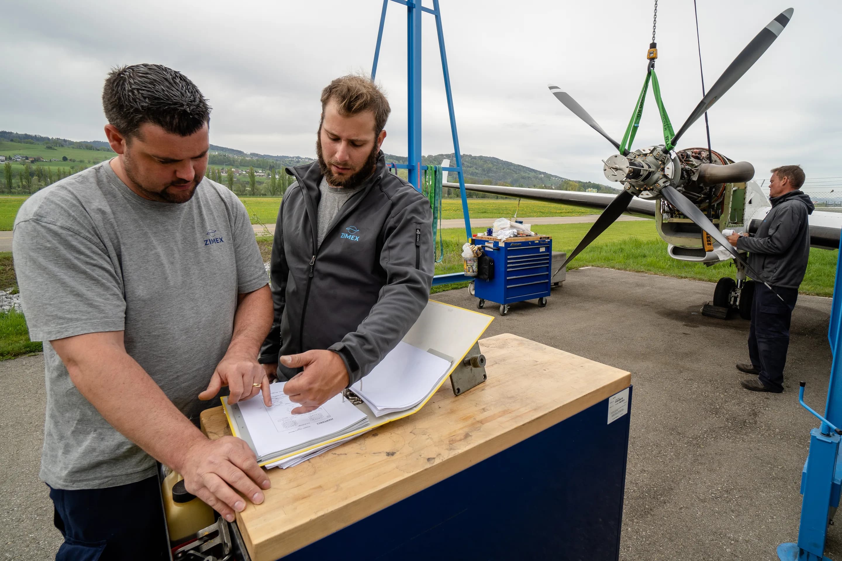 Two Zimex engineers going through paperwork at a workbench at Altenrhein, with a Twin Otter propeller on a hoist behind them.