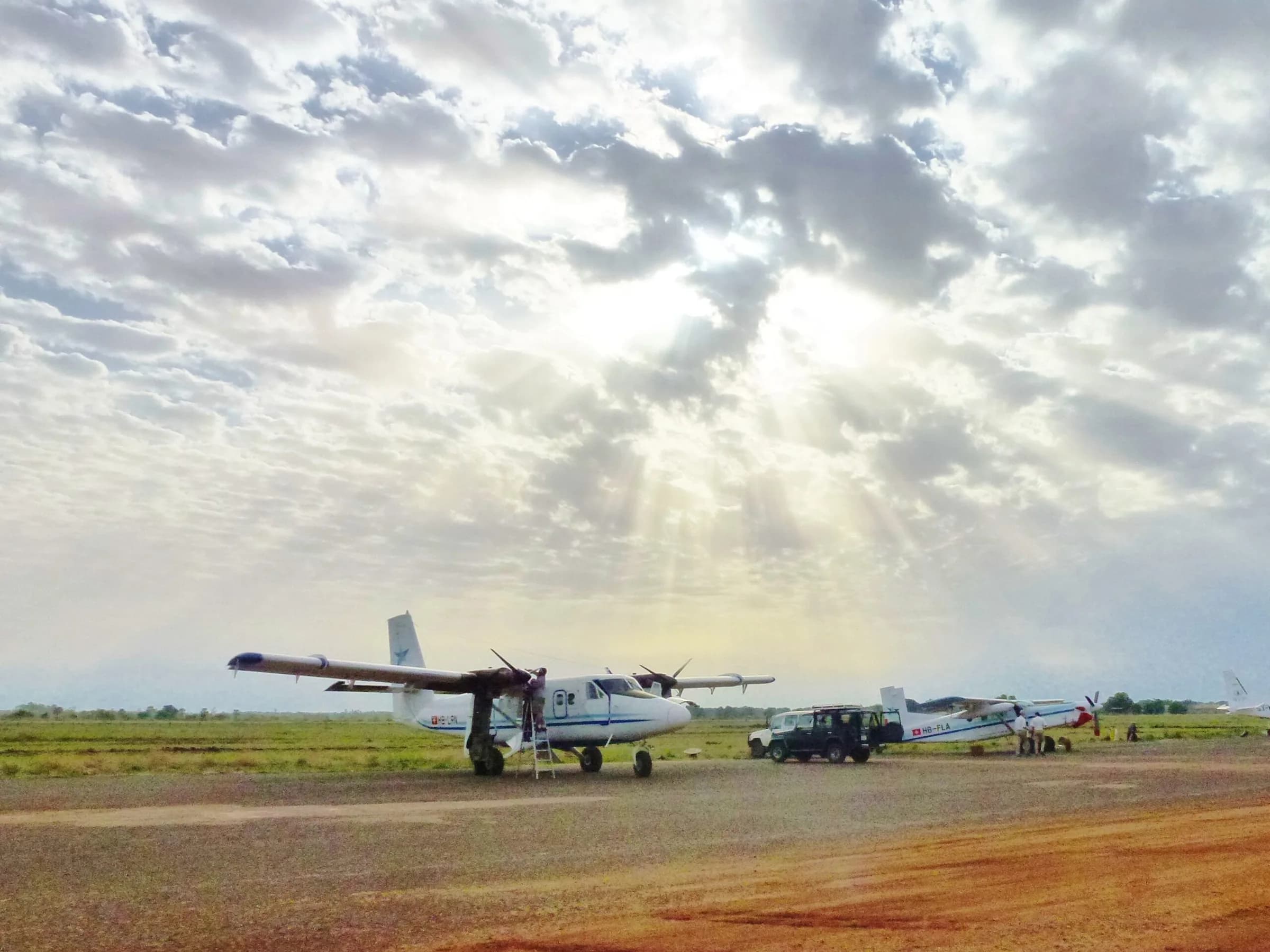 Two Zimex Twin Otters parked on a grass and red-earth strip under sun rays breaking through cloud
