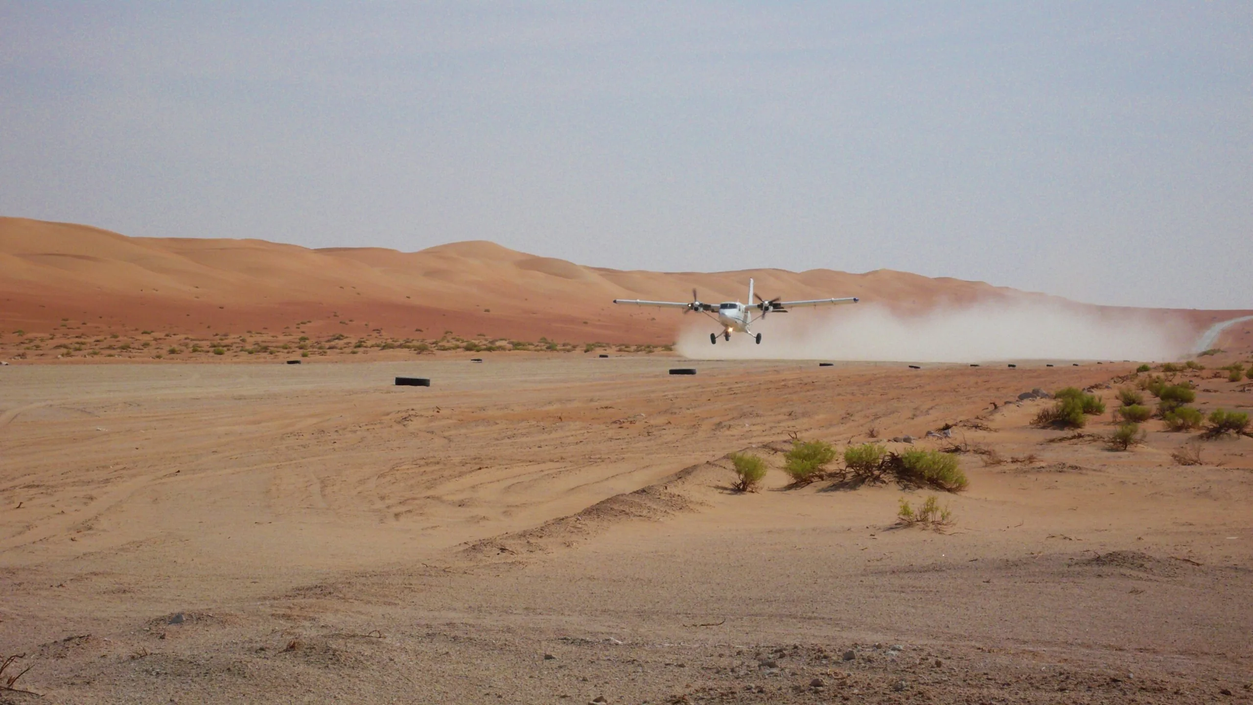 A Zimex Twin Otter taking off from a desert strip, trailing a cloud of dust in front of red dunes