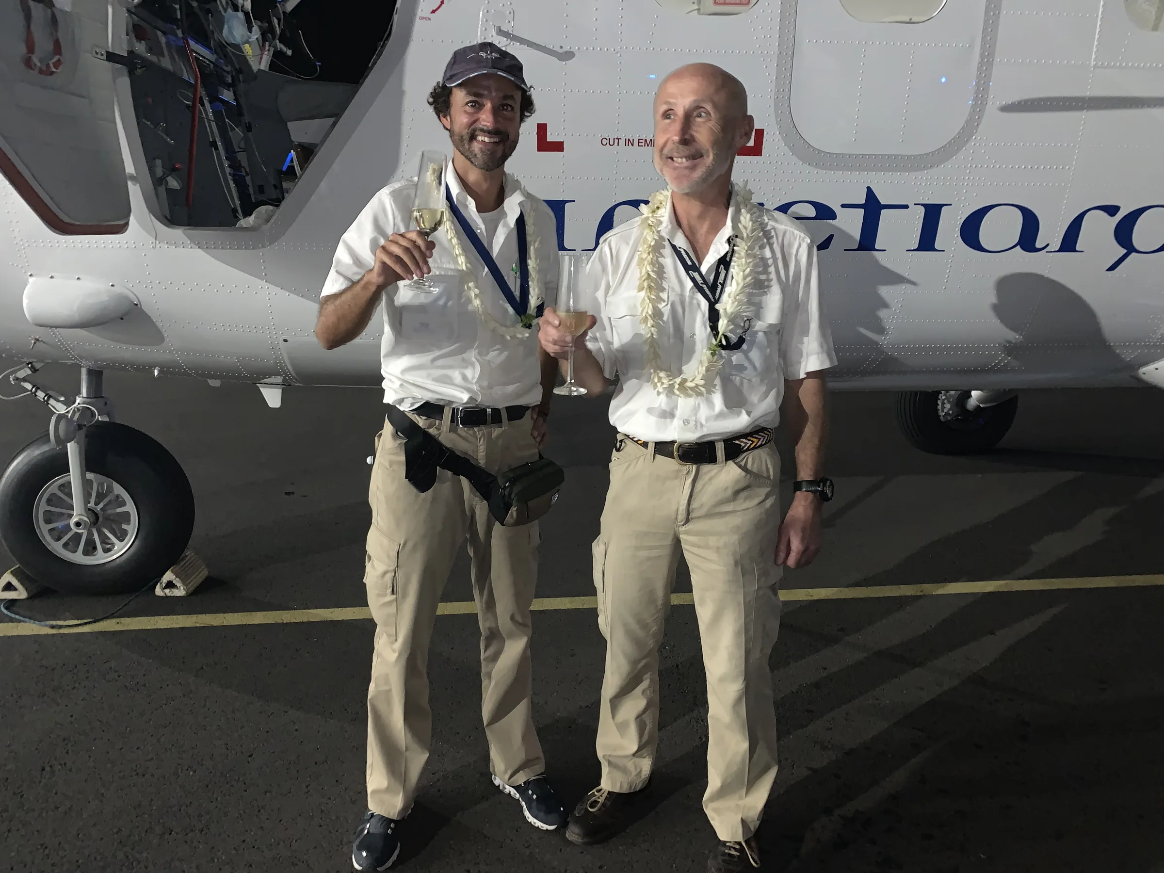Two crew members with flower garlands and glasses in hand in front of the Twin Otter after arrival