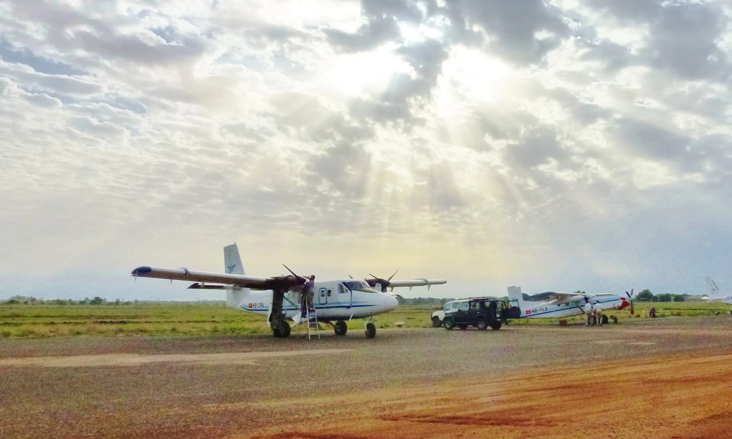 Two Twin Otters and a Land Cruiser on a gravel strip under a cloudy sky